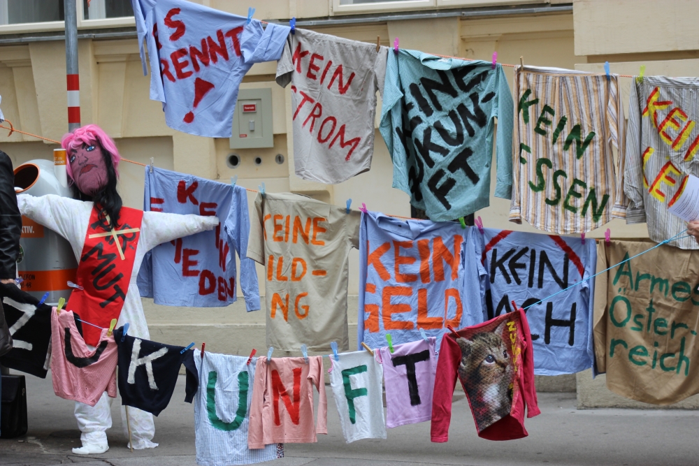 Auf drei Wäscheleinen hängen T-Shirts mit Aufschriften (wie kein Strom, kein Essen, kein Geld) und ein Schriftzug Zukunft
