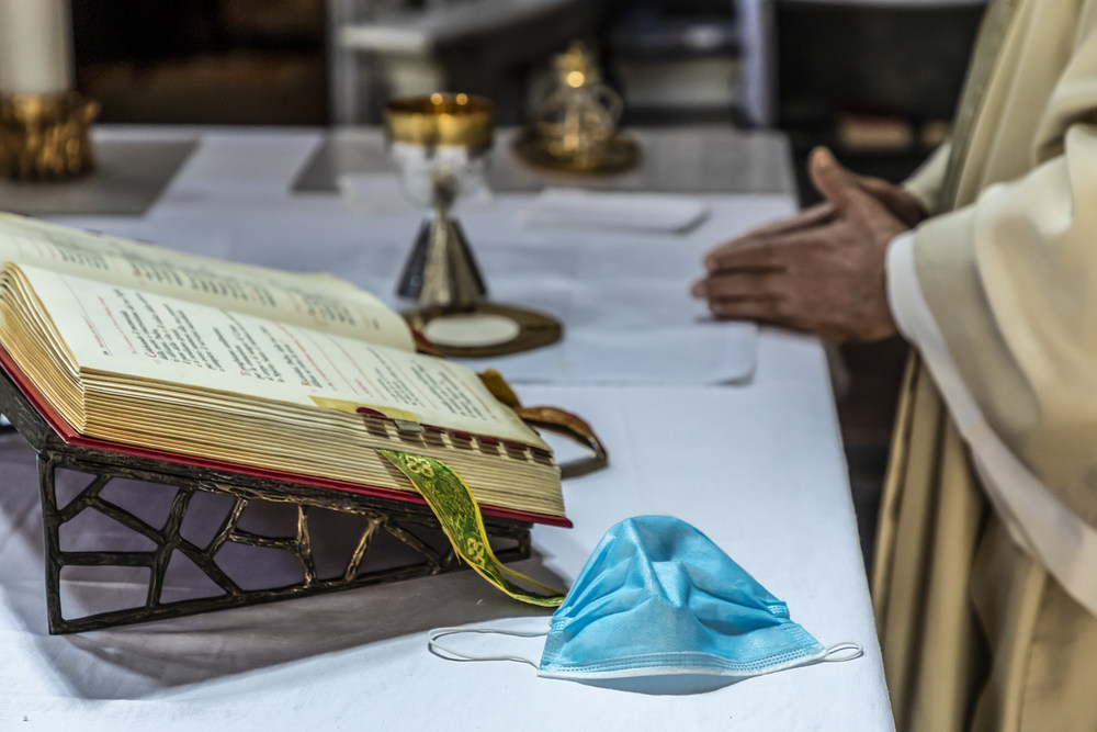 Mundschutz auf dem Altar / Copyright 2020, KNA GmbH, www.kna.de, All Rights Reserved, Stefano Dal Pozzolo Ein Priester faltet die Hände und feiert den Gottesdienst am Altar, auf dem das Messbuch aufgeschlagen liegt, daneben eine einfache Atemschutzmaske, dahinter liturgische Geräte, am 8. Mai 2020 in Rom.